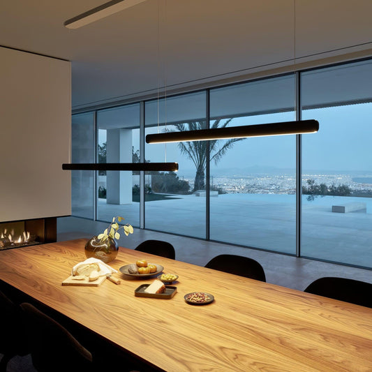 Black linear pendant above a long wood table beside floor-to-ceiling windows